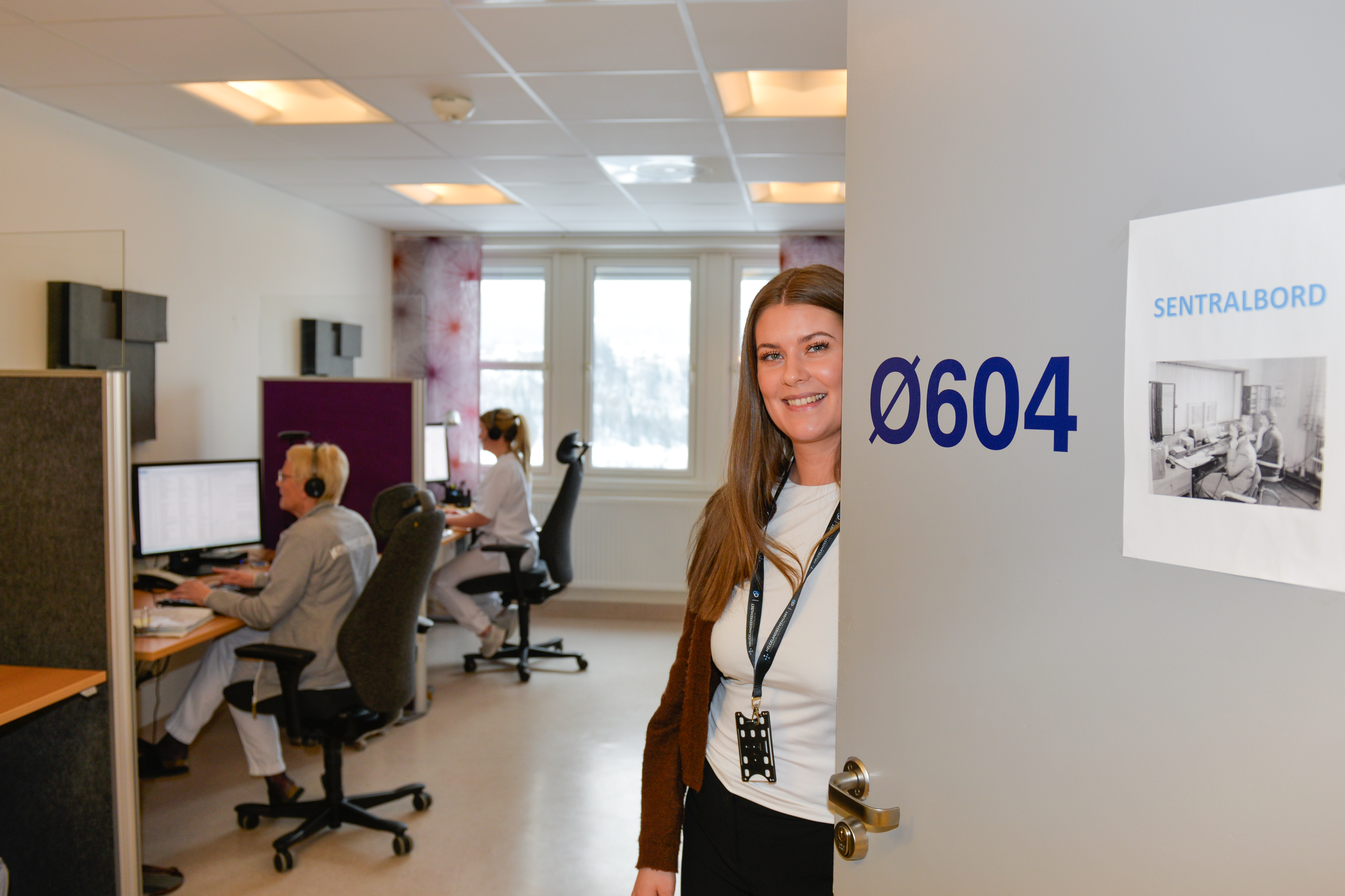 Dame står i en døråpning. I bakgrunnen sitter det flere folk og jobber foran pc-en.