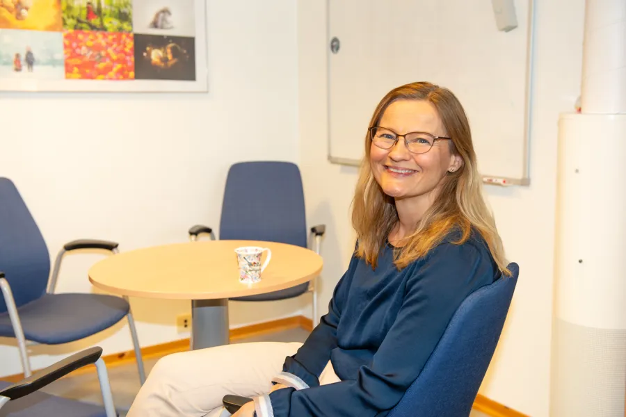 Dame i blå genser sitter i en stol og smiler mot kamera