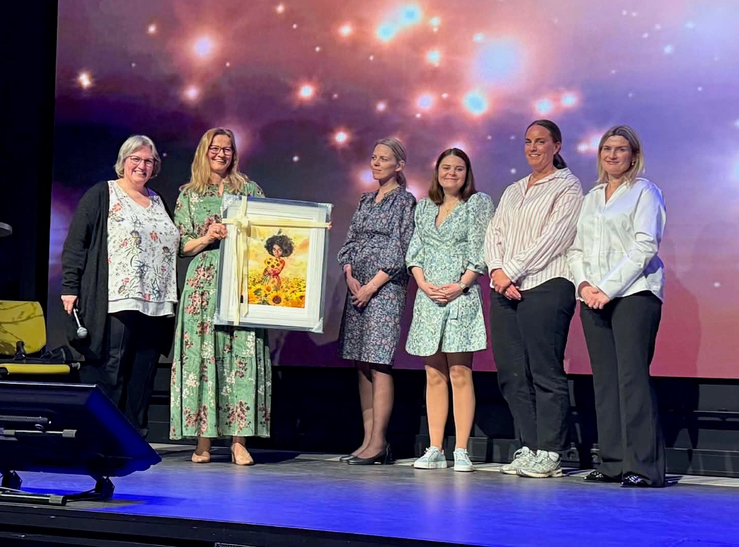 En gruppe mennesker som står på scenen i forbindelse med en prisutdeling. Den ene dama holder et maleri.