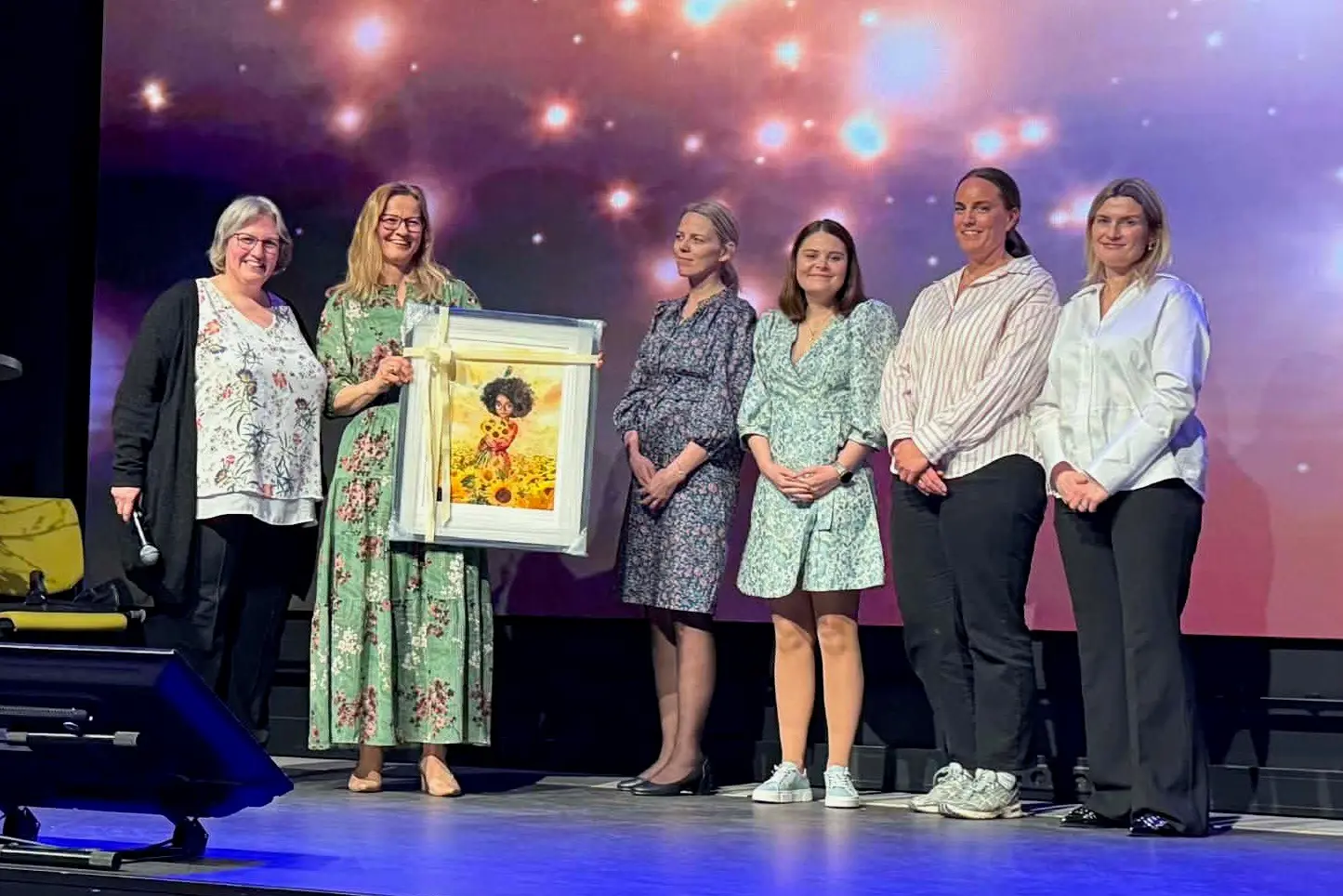 En gruppe mennesker som står på scenen i forbindelse med en prisutdeling. Den ene dama holder et maleri.