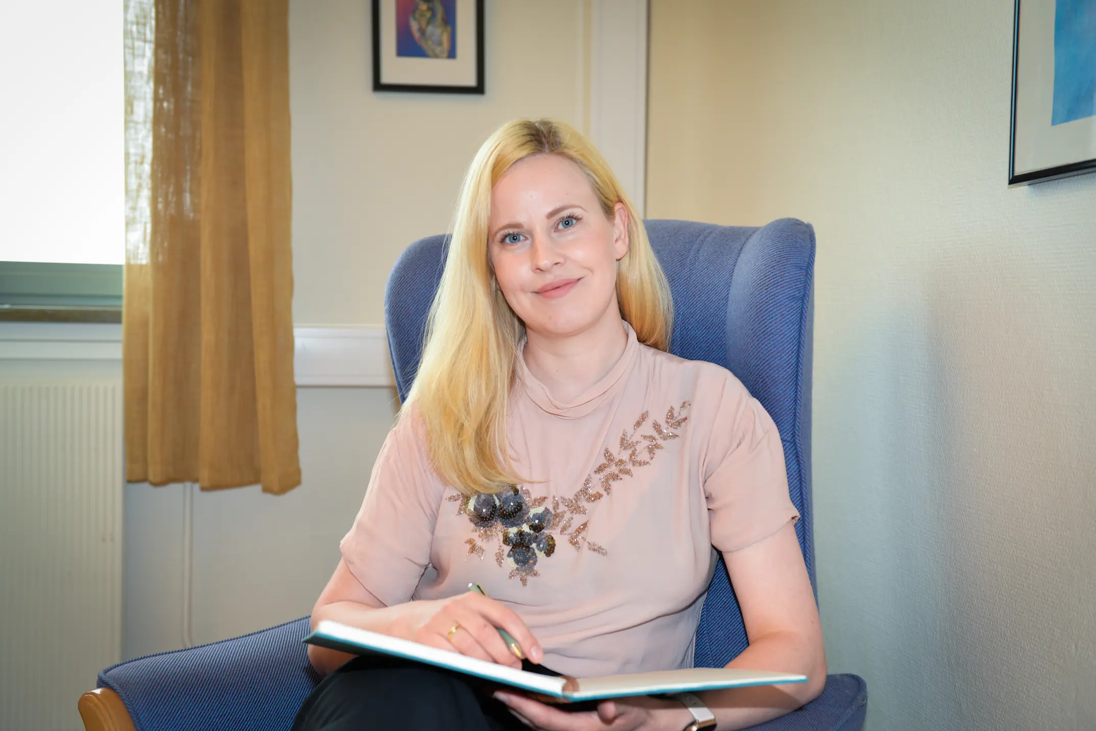 Dame med lyst, langt hår sitter i en blå stål og holder i ei oppslått notatbok