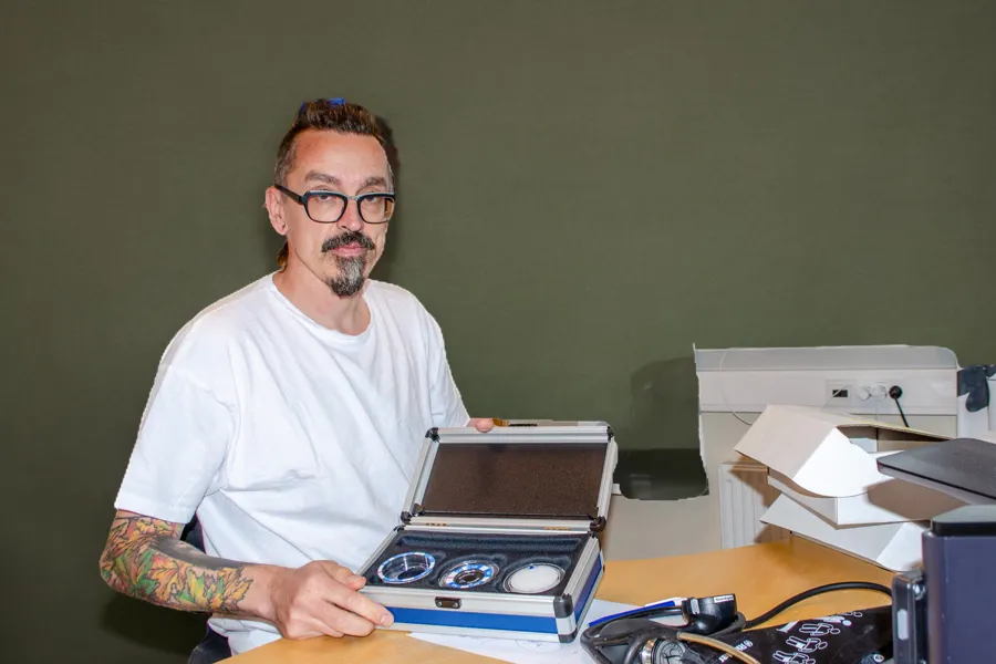 En mann med skjegg som sitter ved et skrivebord med en bærbar datamaskin og annet elektronisk utstyr