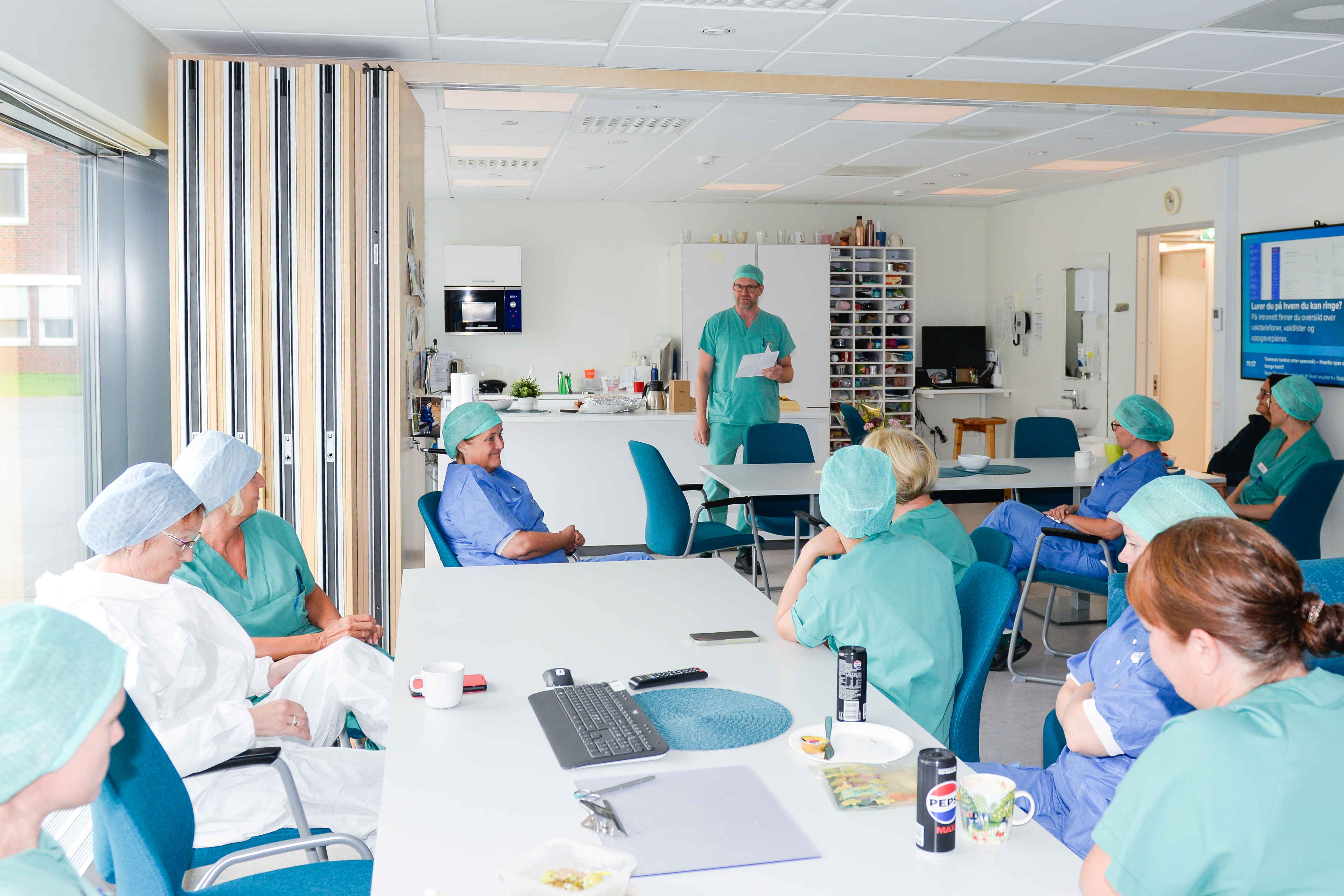 Mange personer sitter på et pauserom. Alle har på seg sykehusklær.