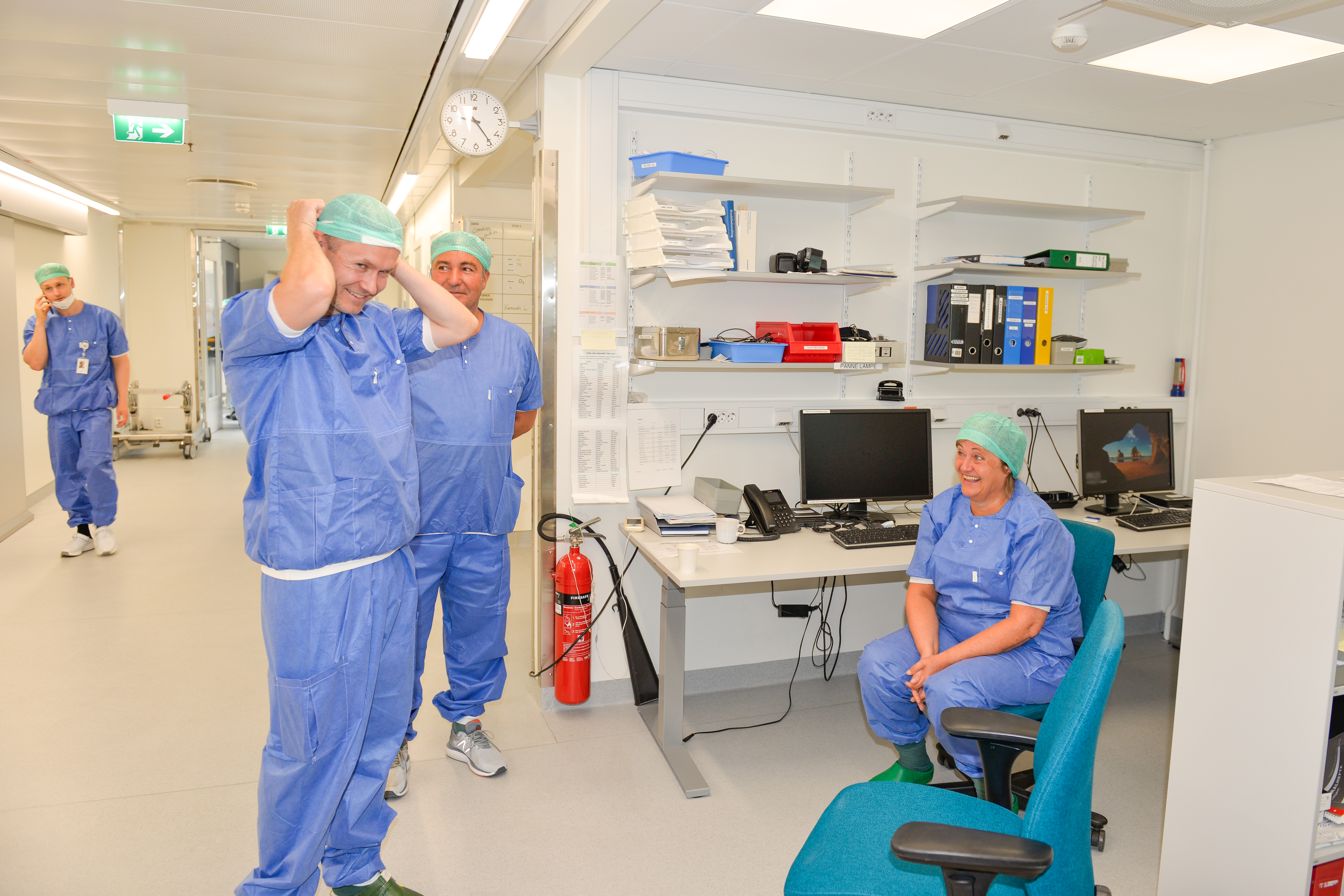 Tre sykehuskledte personer snakker sammen og ler. I bakgrunnen går en mann og prater i telefon