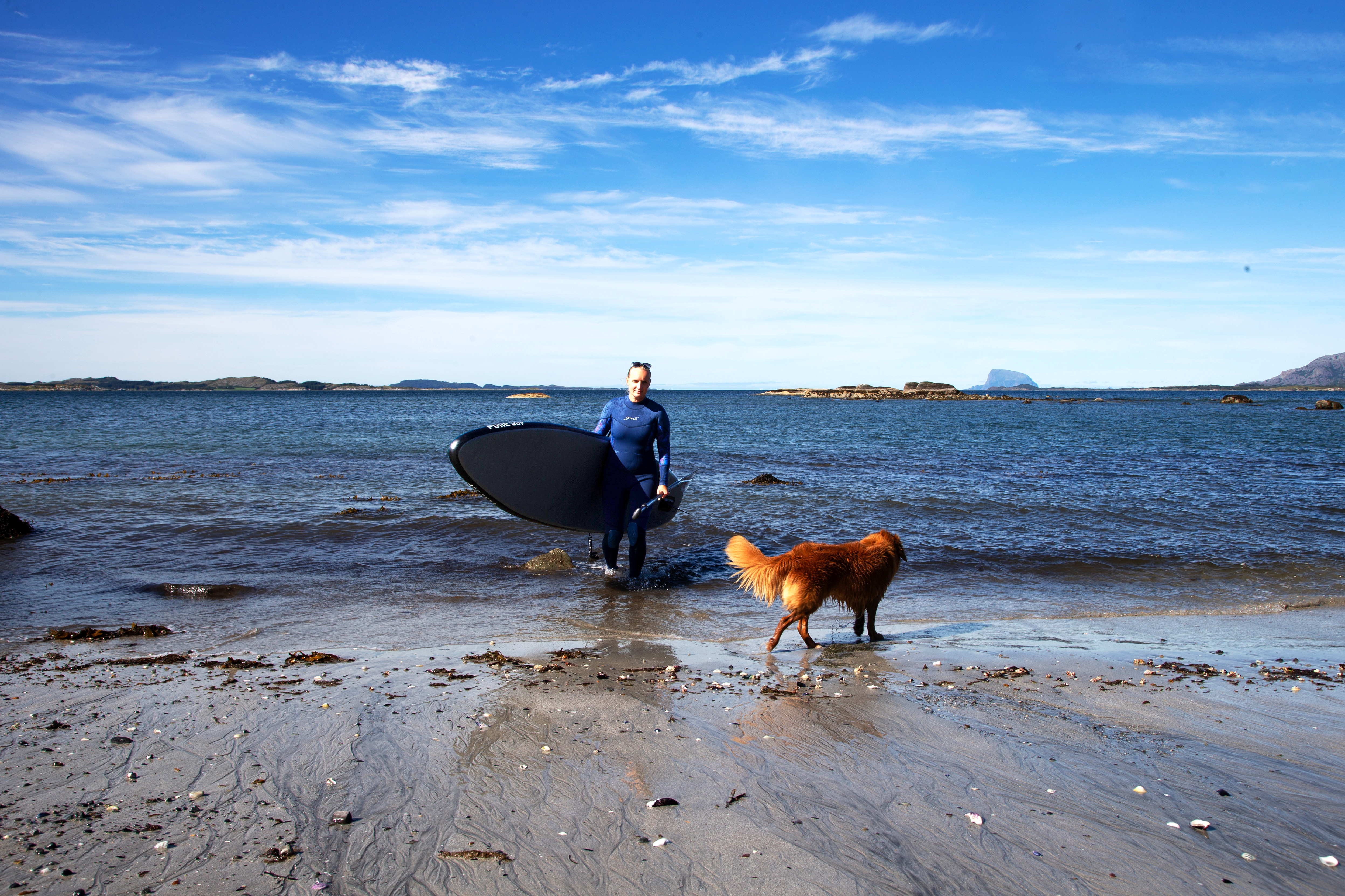 Ei dame kommer gående opp fra havet med et surfebrett under armen