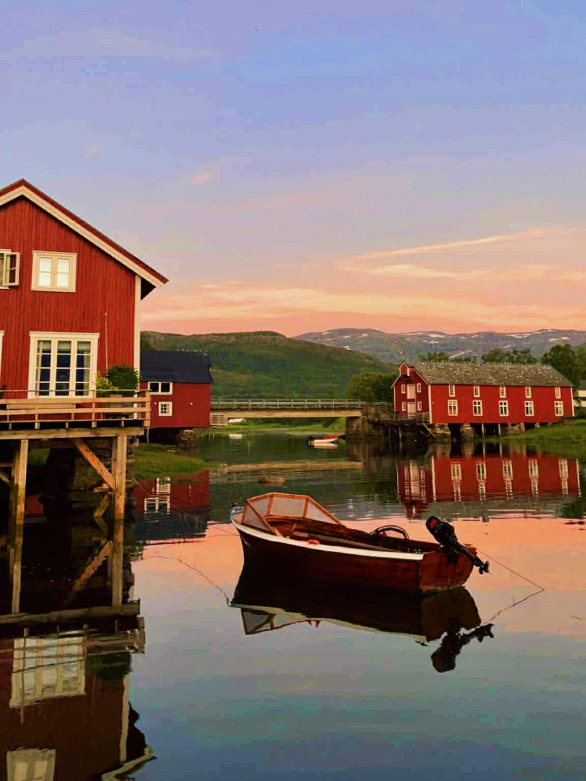 En båt ligger på blikkstille hav med røde sjøhus bak og rosa himmel