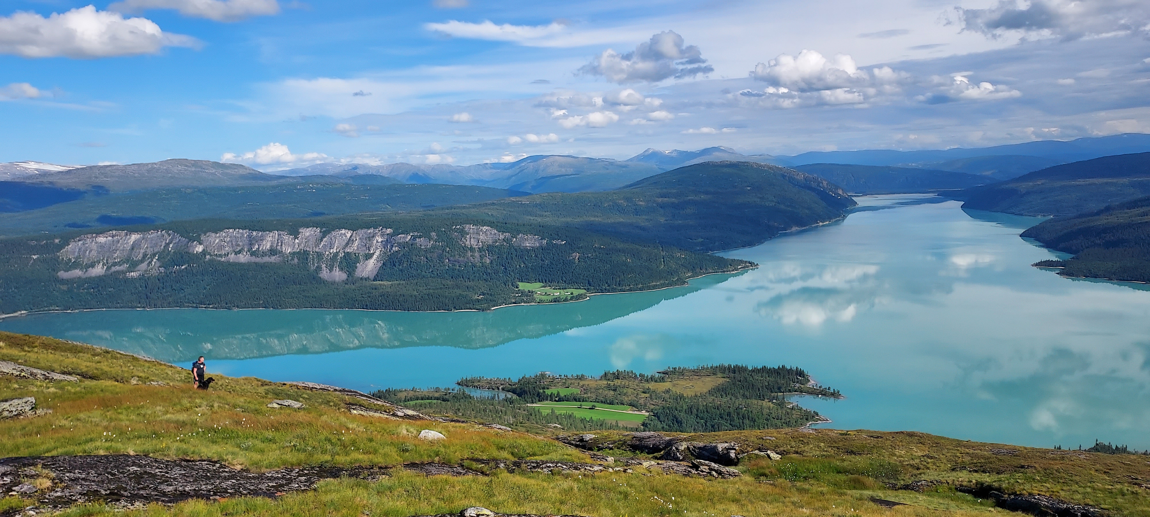 Et oversiktsbilde over naturen med et blått brevann som strekker seg ut, og en person som går oppover mot kameraet