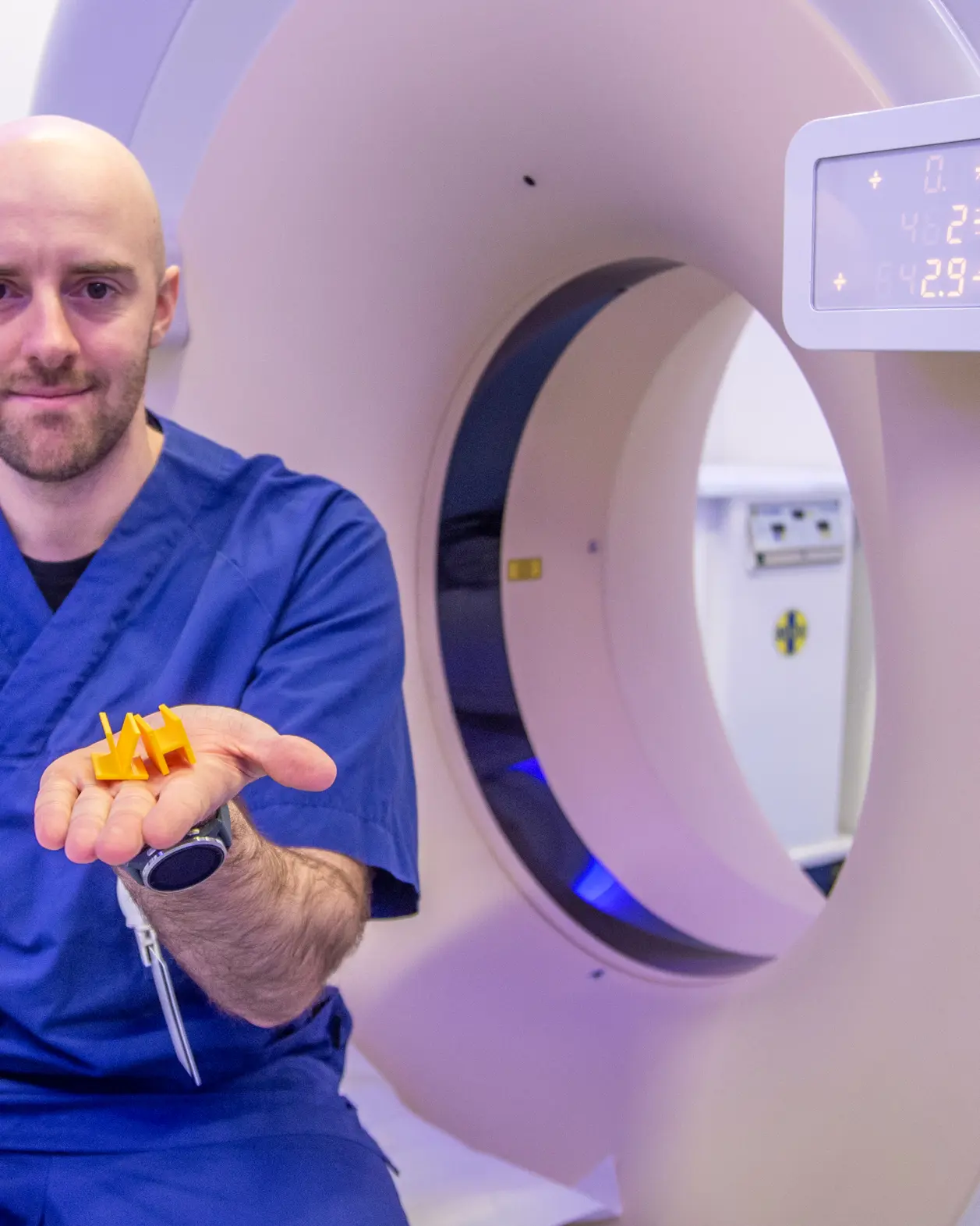 U:ne mann i blå kjeledress som holder gule bokstaver foren en CT-maskin