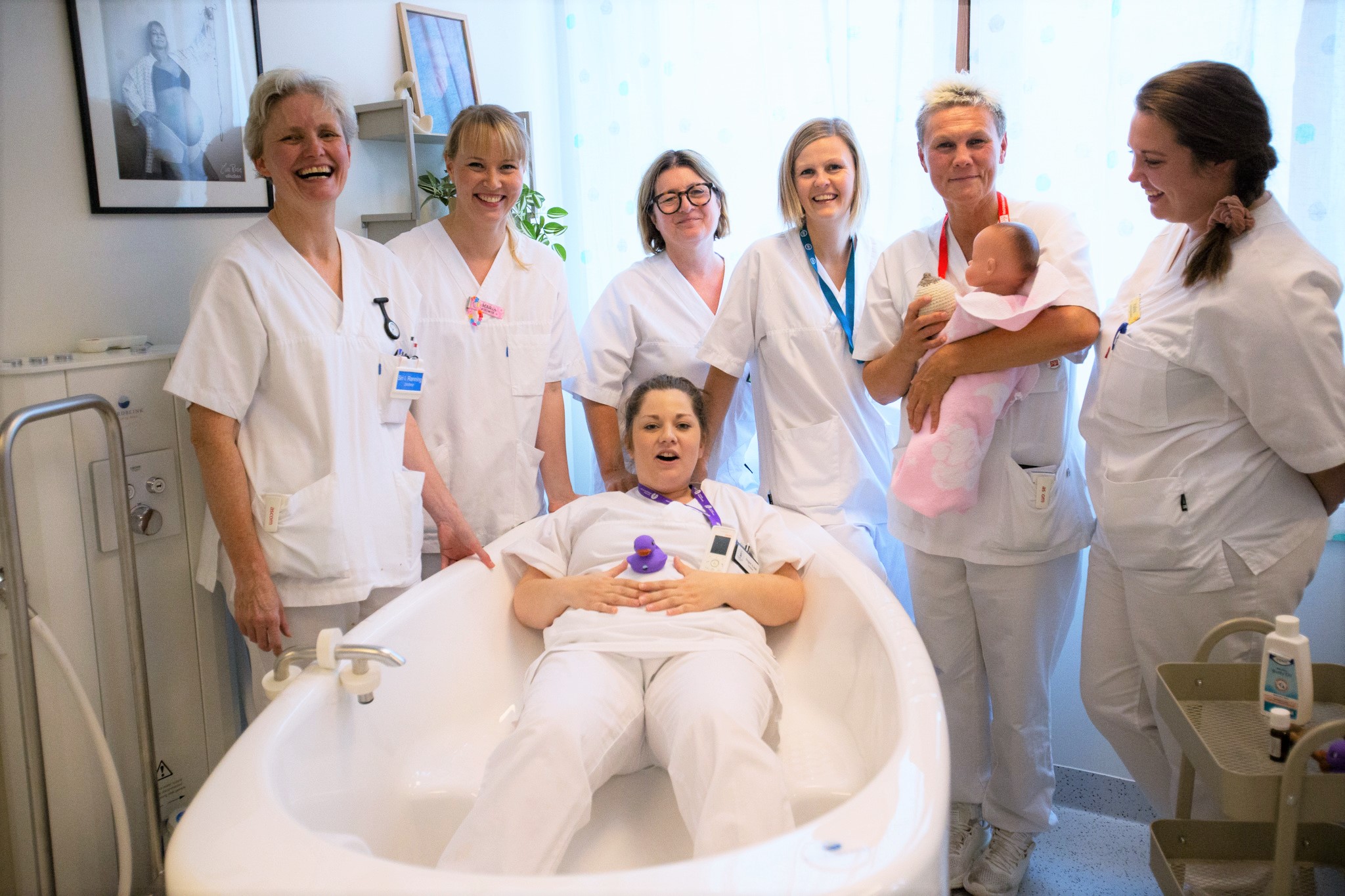 Mange damer står rundt et badekar. I badekaret ligger en dame med ei lekeand på magen.