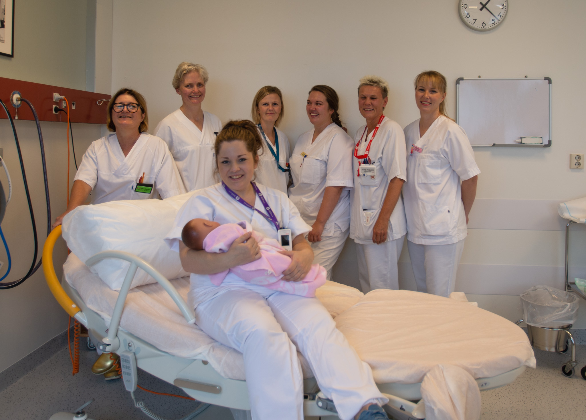 Dame sitter med baby på fanget mens seks damer står rundt henne.