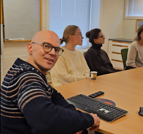 Inne på et møterom sitter flere kolleger rundt et bord, lederen snur seg rundt og smiler til kamera