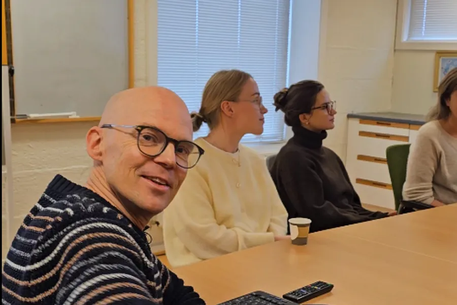 Inne på et møterom sitter flere kolleger rundt et bord, lederen snur seg rundt og smiler til kamera