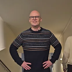 En mann med briller står med armene i siden og smiler til kamera