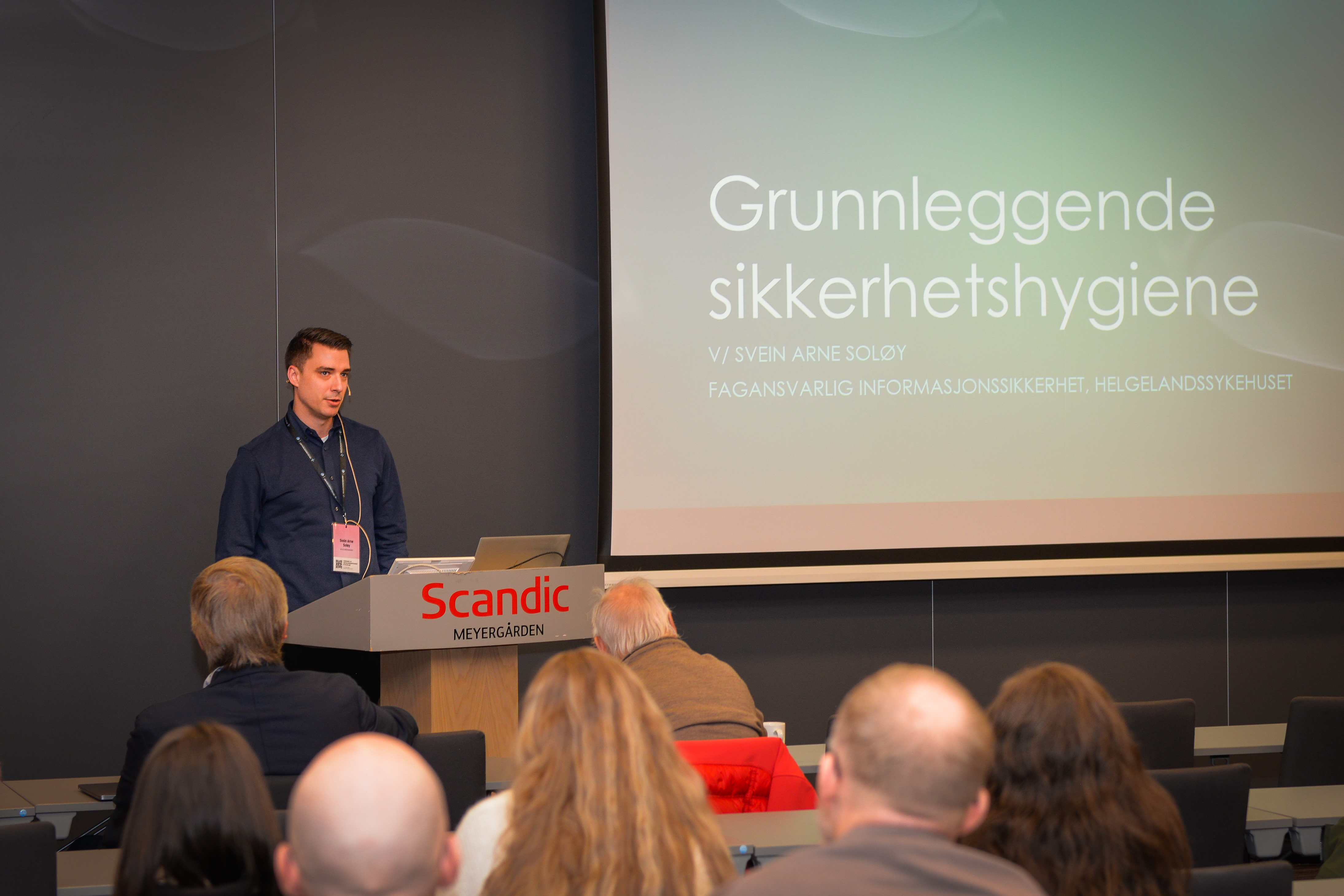 En mann står foran en stor forsamling og snakker om grunnleggende sikkerhetshygiene