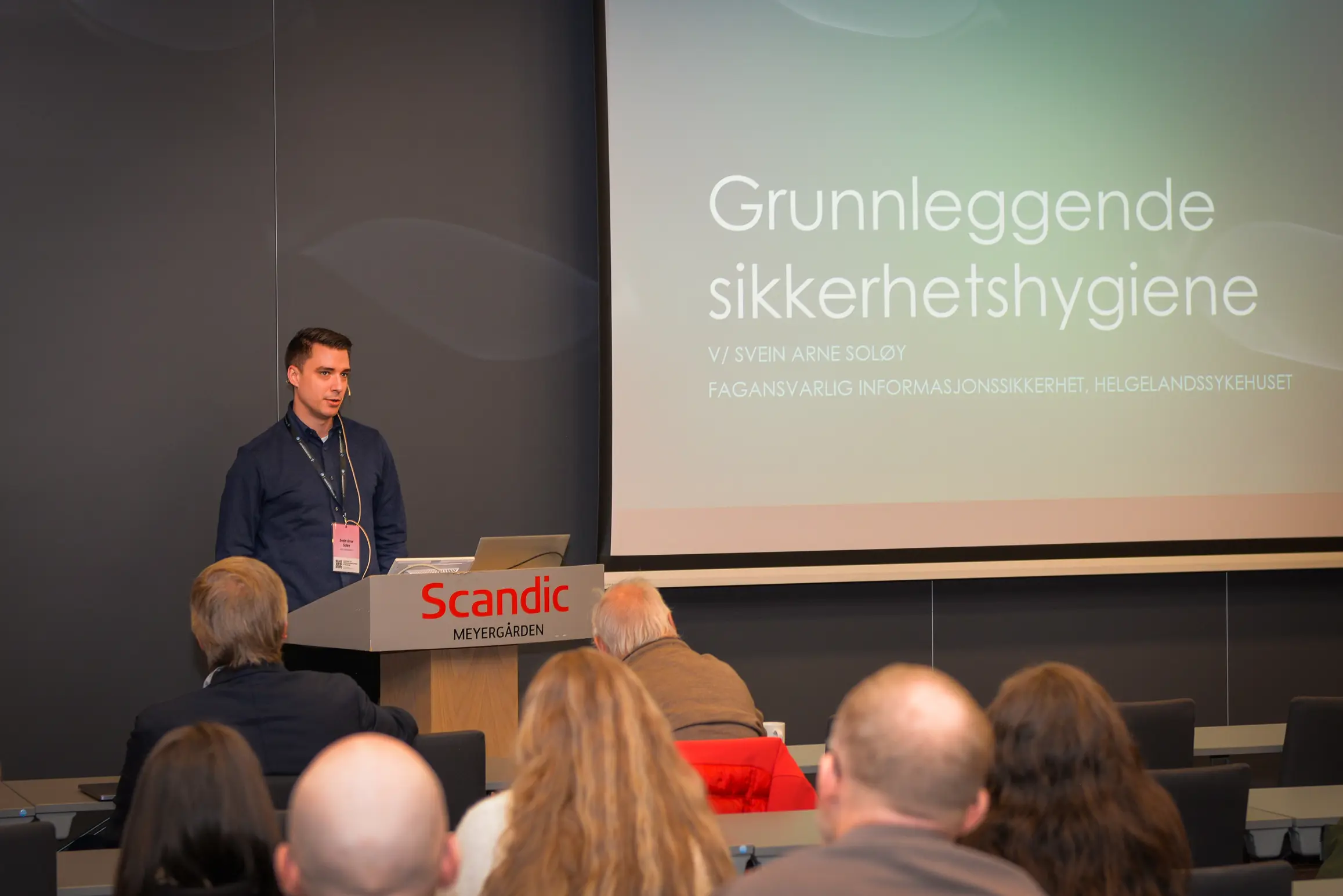 En mann står foran en stor forsamling og snakker om grunnleggende sikkerhetshygiene