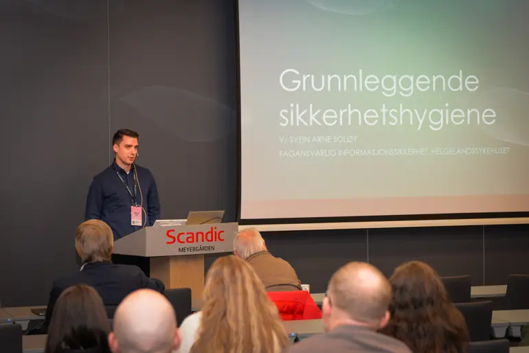 En mann står foran en stor forsamling og snakker om grunnleggende sikkerhetshygiene