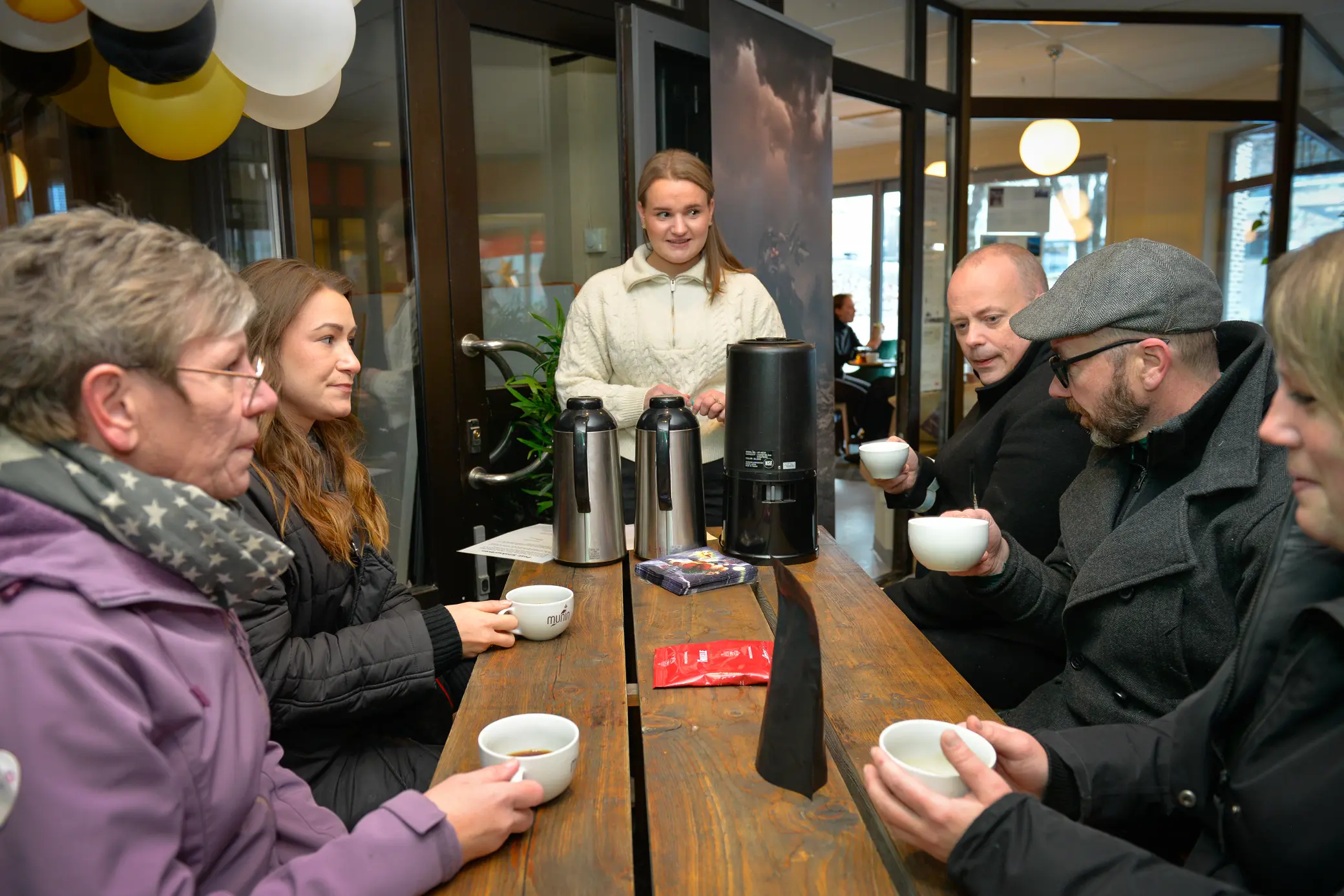 Mange mennesker sitter rundt et bord med hver sin kaffekopp